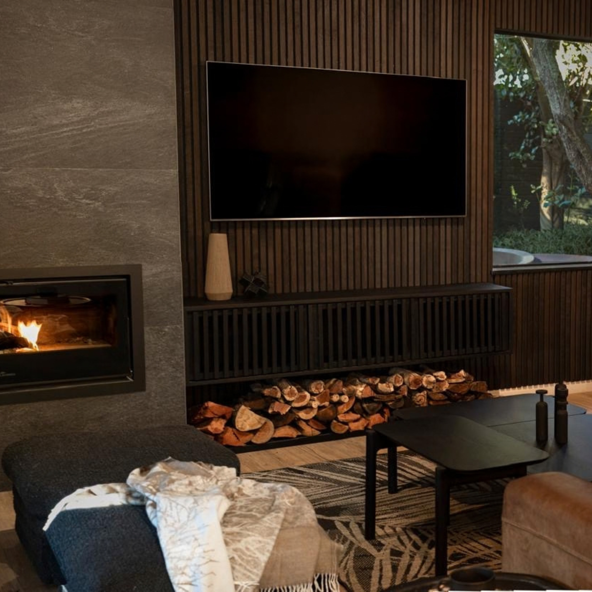 A modern living room interior featuring a wood-burning fireplace with visible logs, a mounted flat-screen TV on acoustic slatted wood wall, a dark console, and a window looking out to trees.