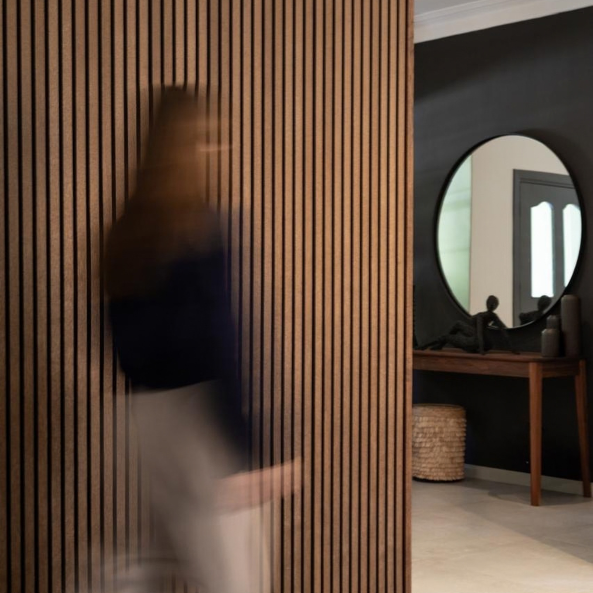 A modern interior scene with tall Luun Acoustic Wall Panels and a blurred person walking in front of them, and a dark wall with a round mirror above a wooden console table.