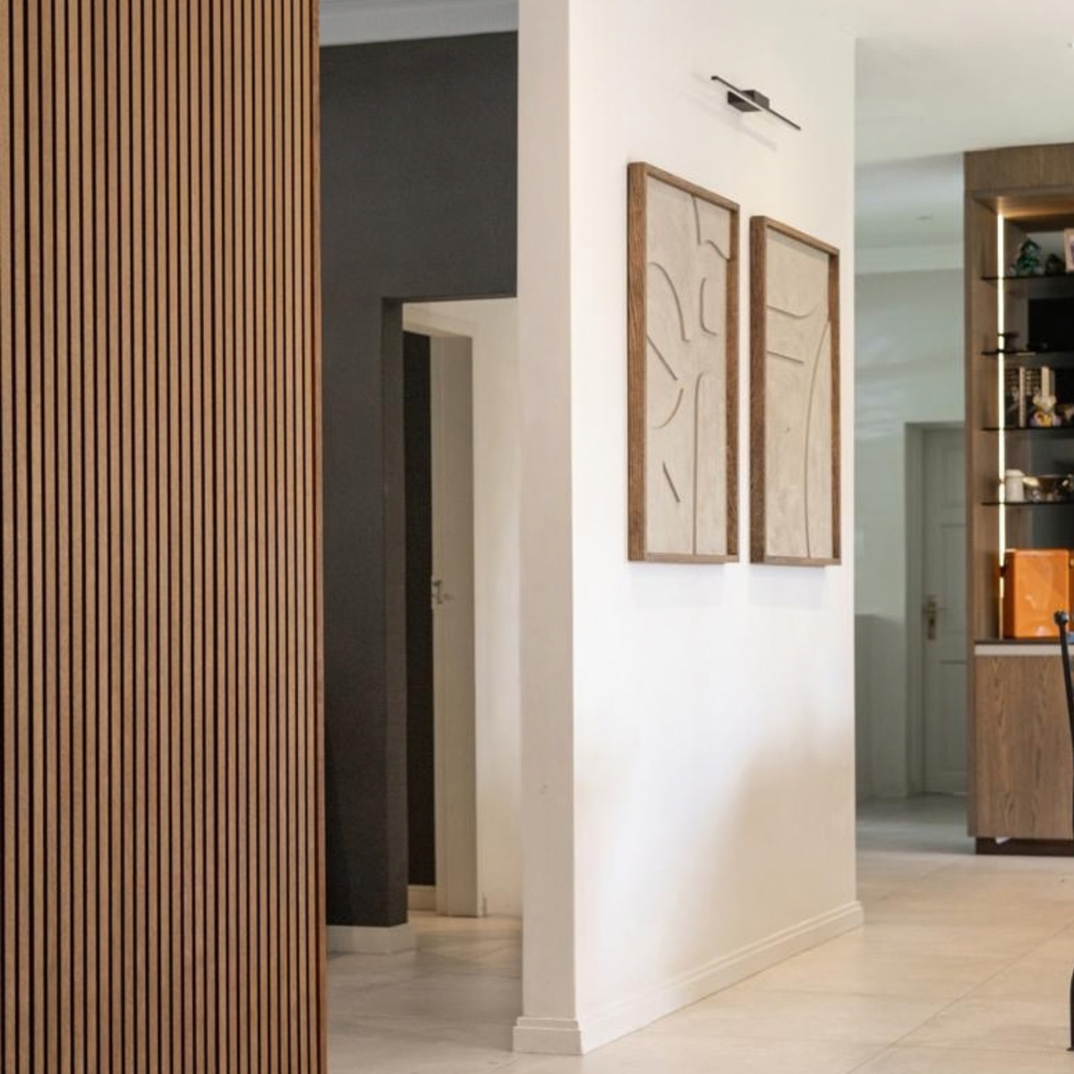 A modern interior hallway with two framed abstract artworks on a white wall, a slatted wood partition on the left, and a built-in shelving unit in the background.